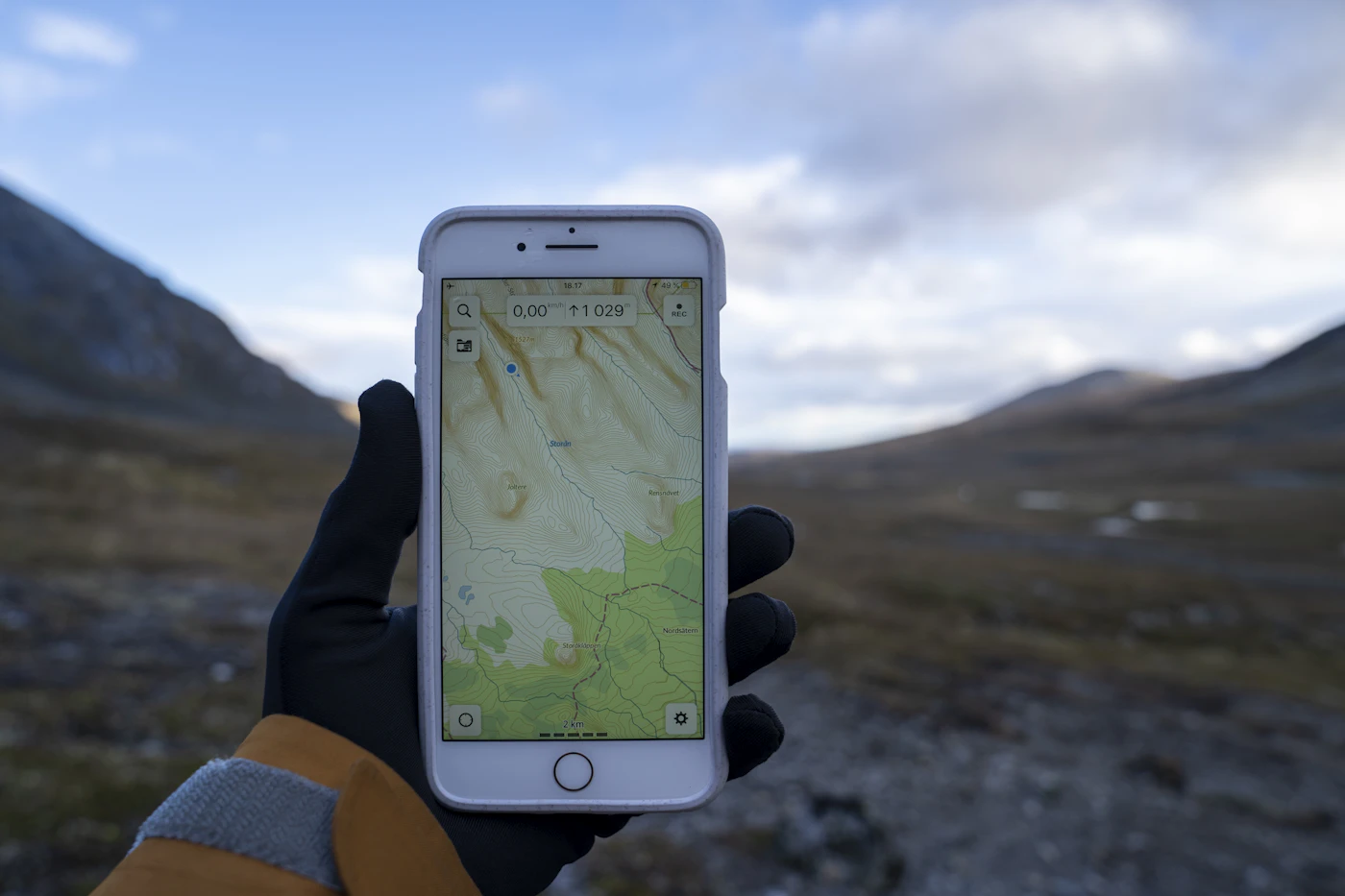 A gloved traveller holding a phone displaying a topographic map in a mountain landscape