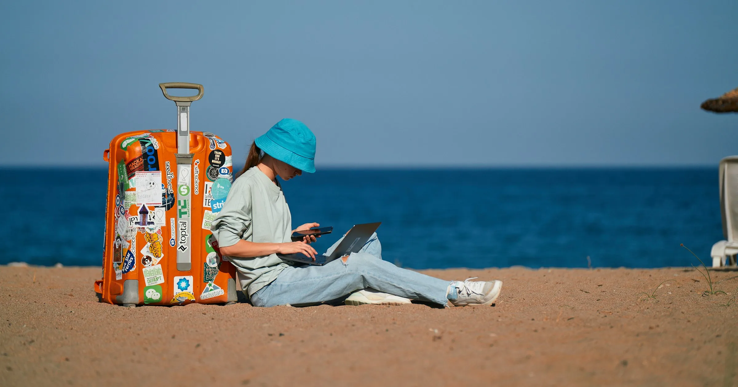 A traveller in a blue bucket hat working on a laptop on a beach beside a suitcase covered in stickers