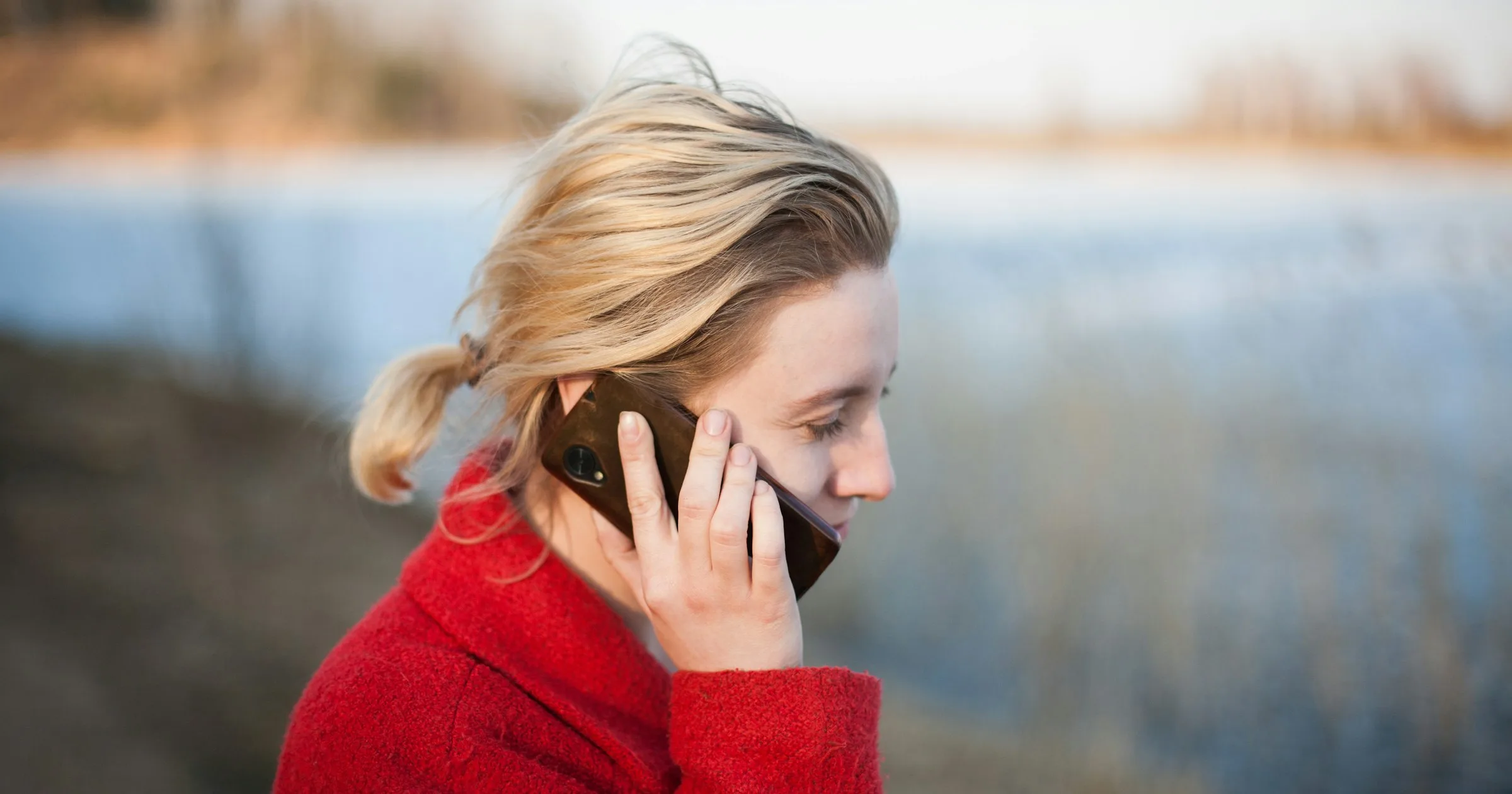 A woman in a red coat making a phone call outdoors by a lake